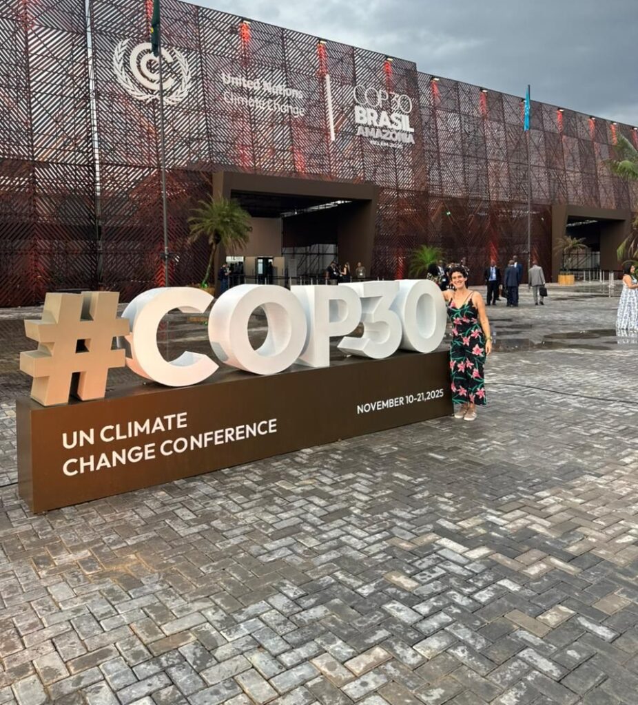 Lari Torello sorri ao lado do letreiro tridimensional da "#COP30", com o grande pavilhão do evento "United Nations Climate Change" ao fundo.