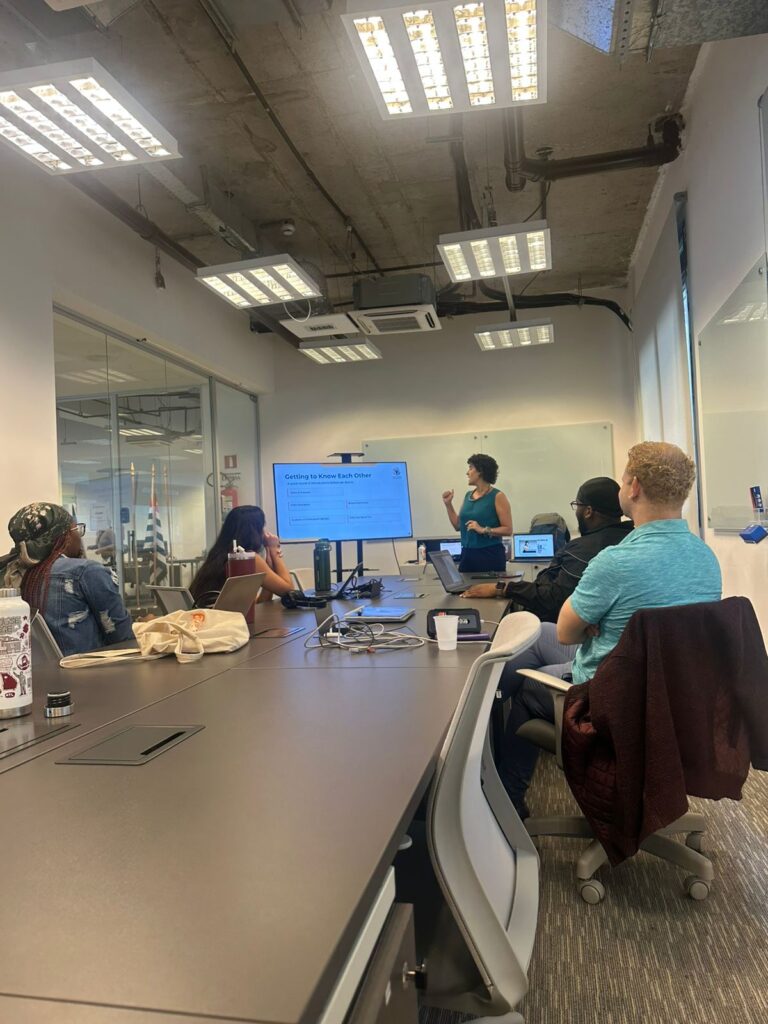 Pessoas sentadas ao redor de uma mesa retangular em uma sala de reuniões moderna, acompanhando a apresentação de uma mulher ao fundo, em pé ao lado de uma tela.