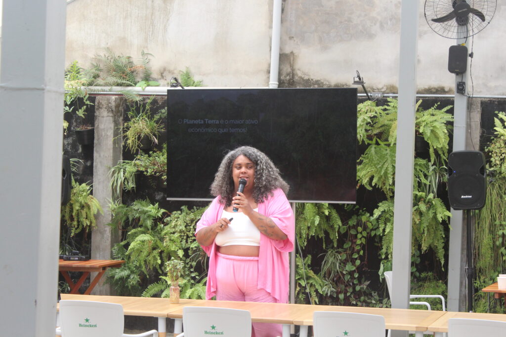 Mulher negra de cabelo afro volumoso, vestindo blazer e calça rosa com top branco, fala ao microfone durante apresentação. Ao fundo, há um painel de LED desligado sobre uma parede de vegetação vertical com folhagens verdes.