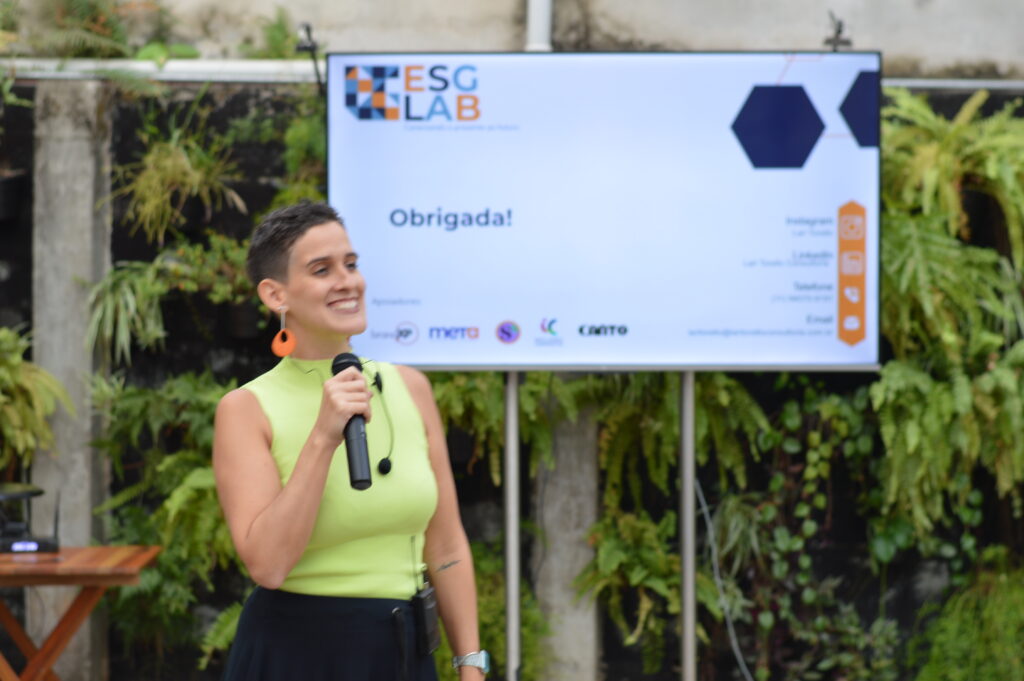 Lari Torello, de regata verde-lima, segura um microfone enquanto fala no palco, sorrindo. Ao fundo, o painel do evento "ESG Lab" exibe a mensagem "Obrigada!" à frente de uma parede coberta por folhagens.