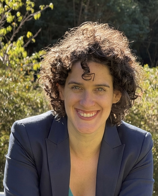 Retrato de Larissa Torello sorrindo, com cabelos cacheados, usando blazer escuro e ao ar livre.