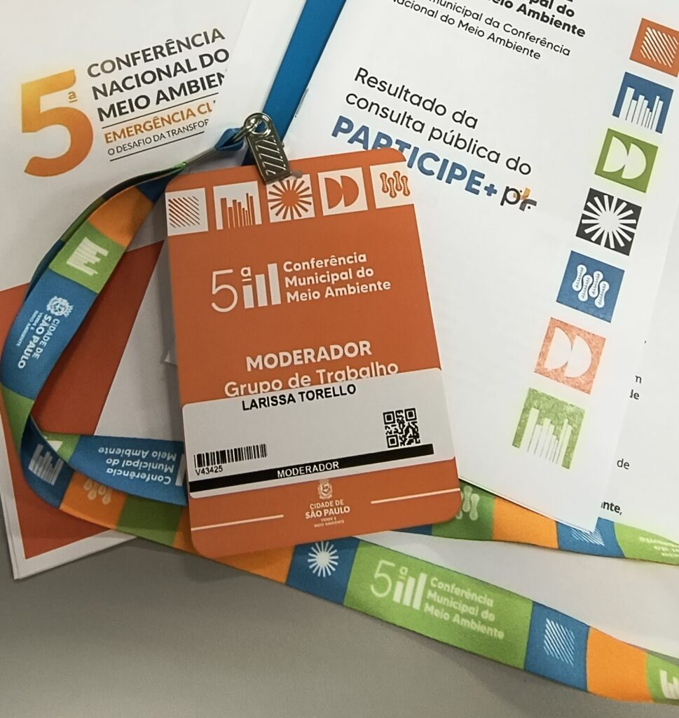 Crachá laranja de moderação do evento "5ª Conferência Municipal do Meio Ambiente" em nome de Larissa Torello, com cordão colorido e documentos do evento ao fundo.