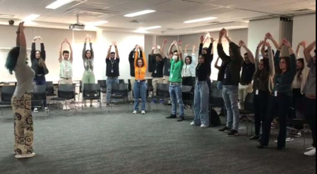Uma instrutora em pé em uma sala de treinamento, de frente para um grupo de participantes dispostos em círculo. Todos os participantes estão com os braços levantados e estendidos acima da cabeça, realizando um exercício de alongamento ou relaxamento durante um treinamento de inteligência emocional.