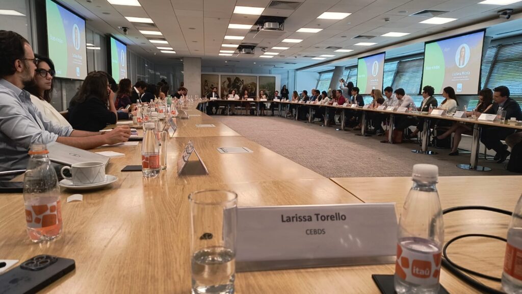 Placa de identificação com o nome Larissa Torello (CEBDS) sobre uma grande mesa retangular em uma sala de reuniões repleta de executivos e autoridades. Ao fundo, telas exibem a apresentação do evento.